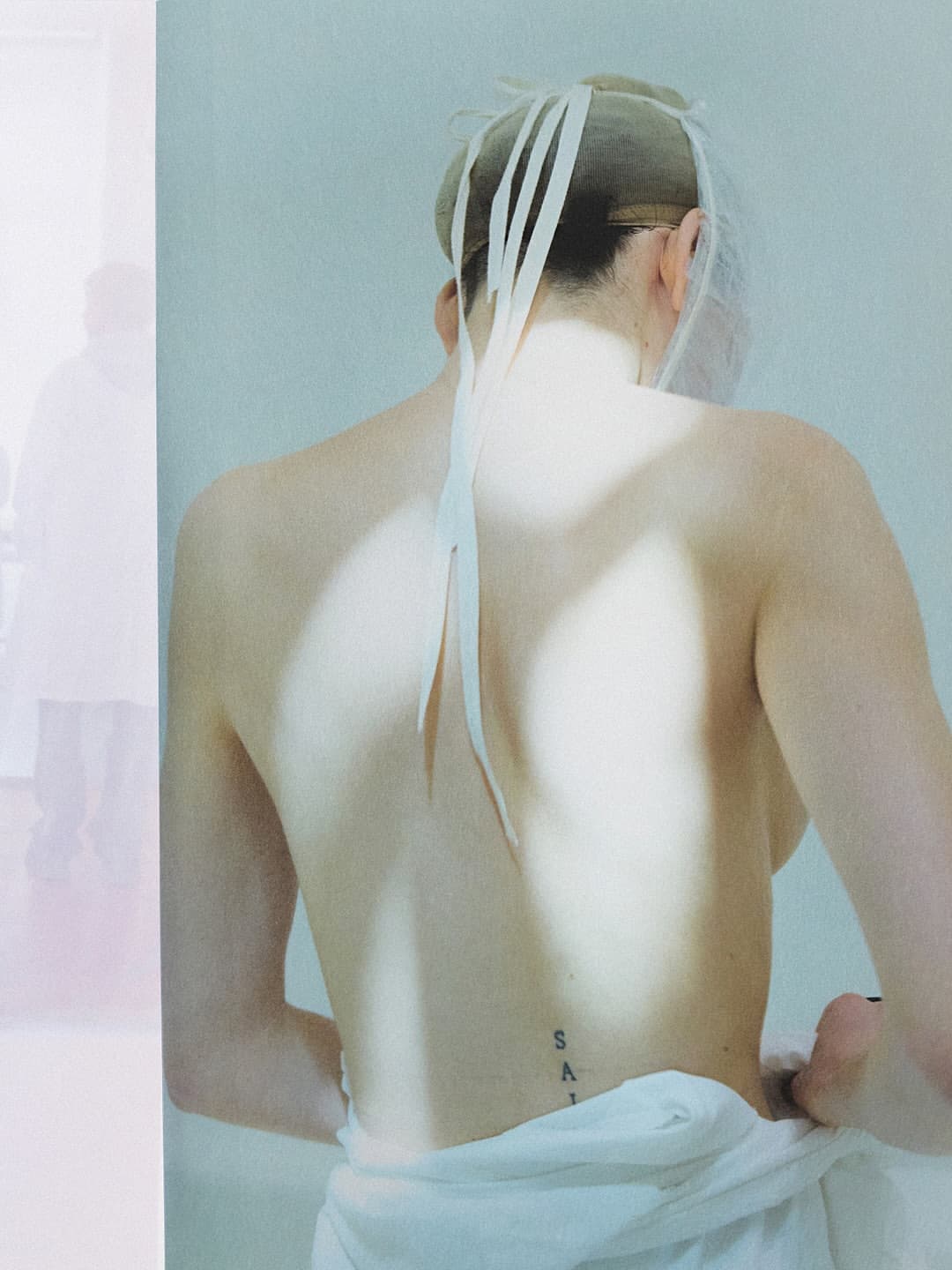 Model seen from behind wearing a hairnet with hanging ribbons and a white garment wrapped around the lower back, against a pale backdrop.