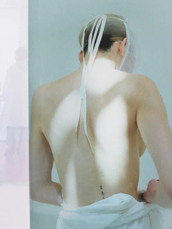 Model seen from behind wearing a hairnet with hanging ribbons and a white garment wrapped around the lower back, against a pale backdrop.