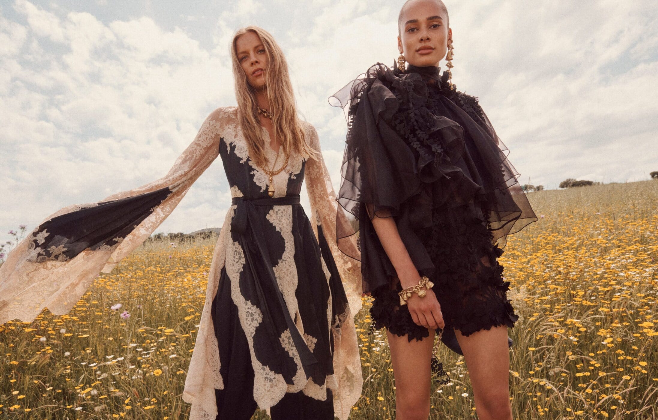 Two female models in black and cream designer dresses standing in a wildflower field under an overcast sky.