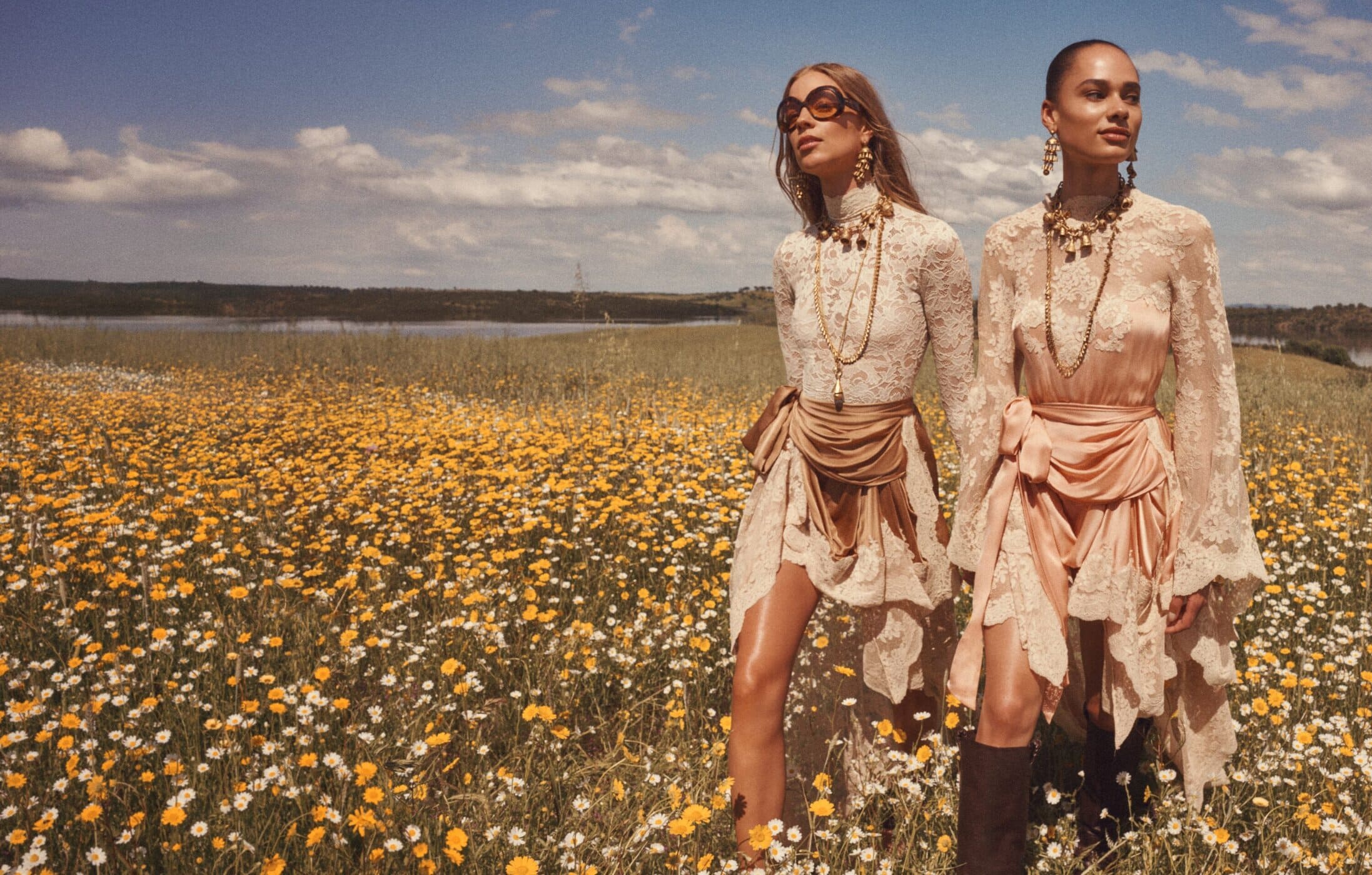 Two female models in lace dresses standing in a wildflower field under a partly cloudy sky for Zimmermann