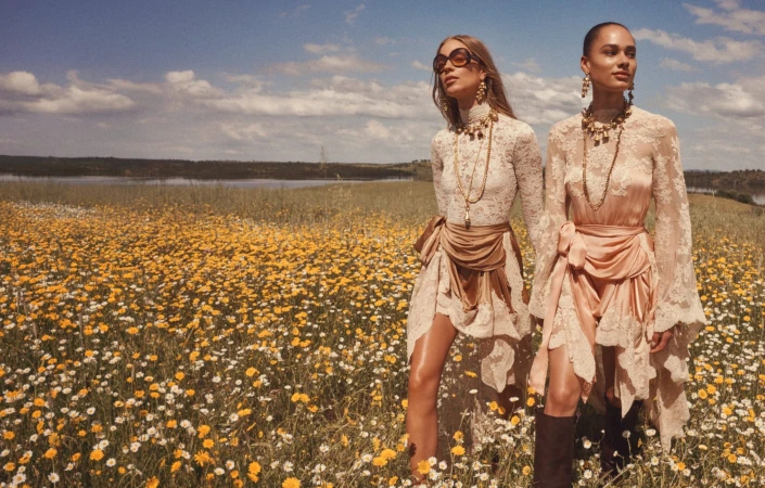 Two female models in lace dresses standing in a wildflower field under a partly cloudy sky for Zimmermann