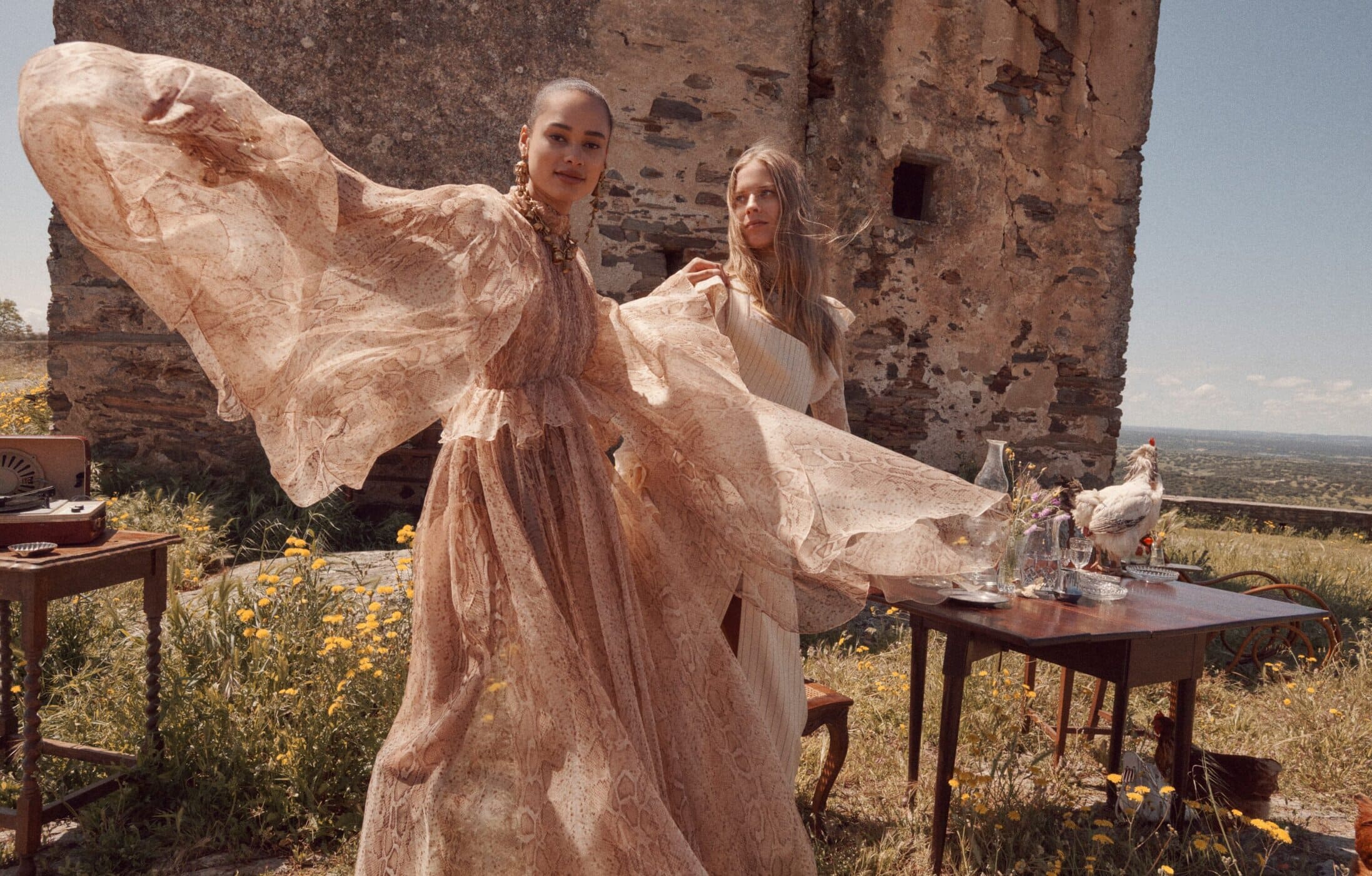Two female models in flowing dresses posing outdoors against an ancient stone wall for Zimmermann Fall 2025 ad campaign.