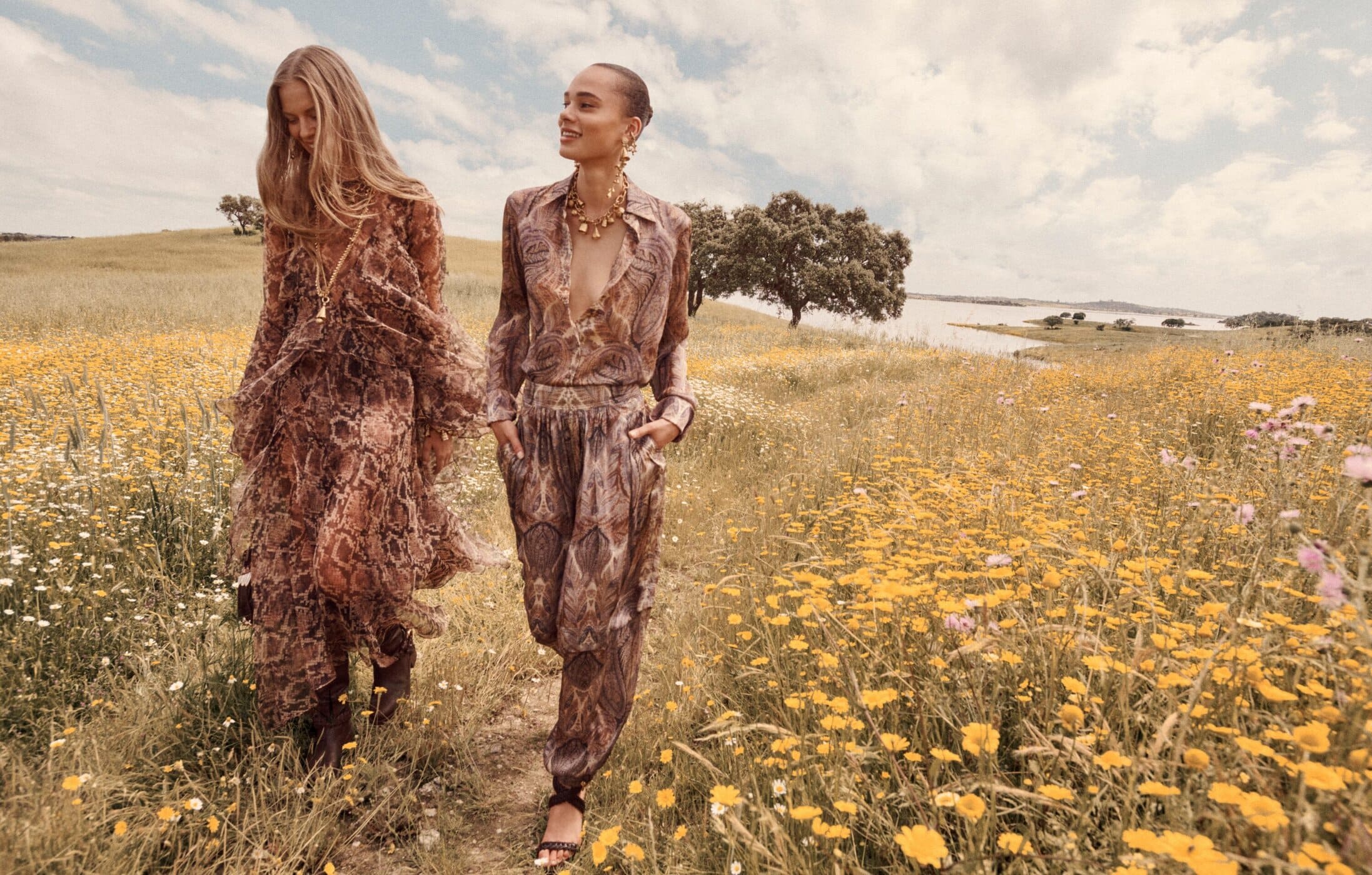 Two female models walking through a grassy field filled with yellow wildflowers under an open cloudy sky.