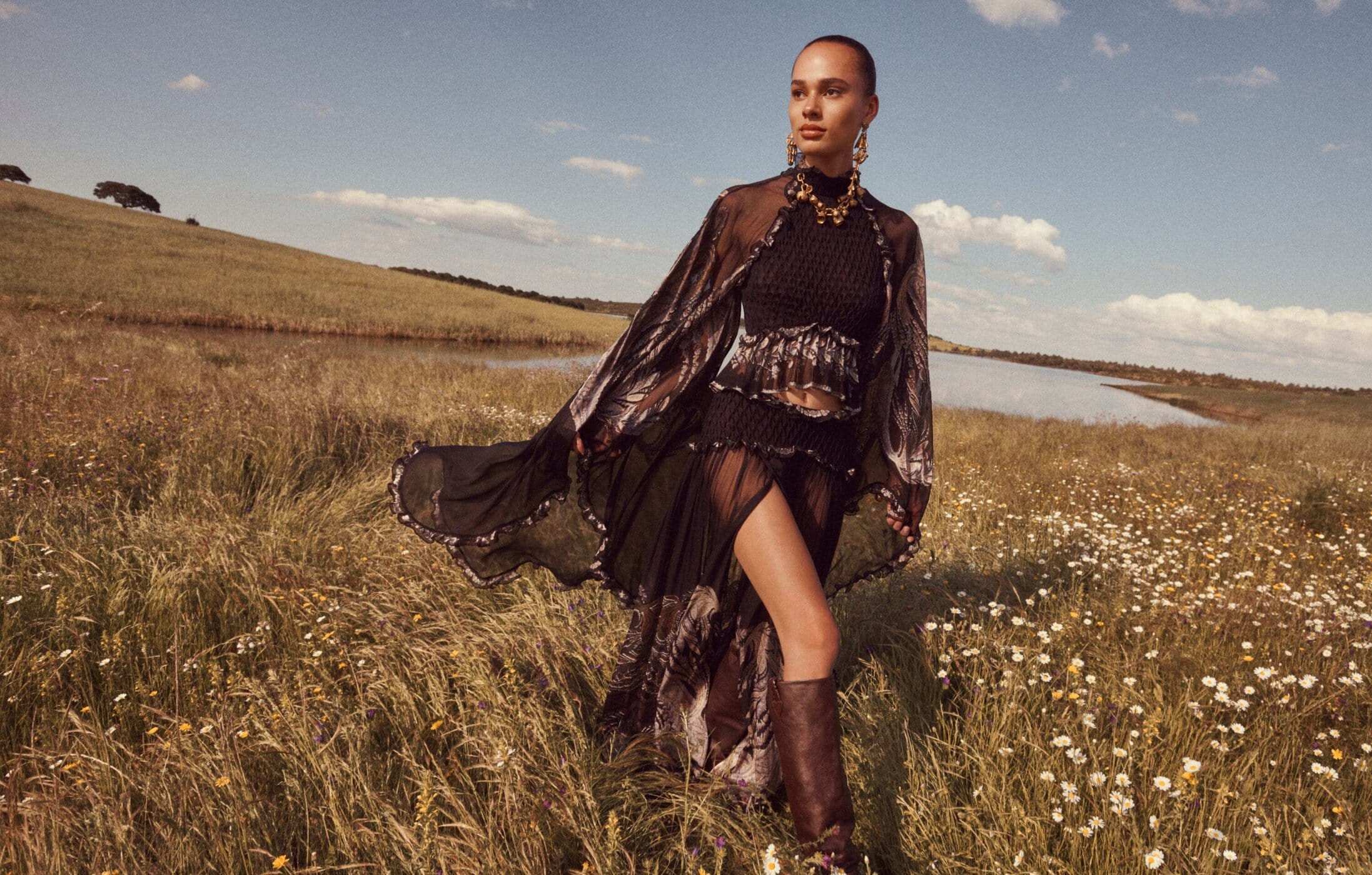 Model in a black sheer dress and boots walking through a grassy meadow for Zimmermann Fall 2025 campaign