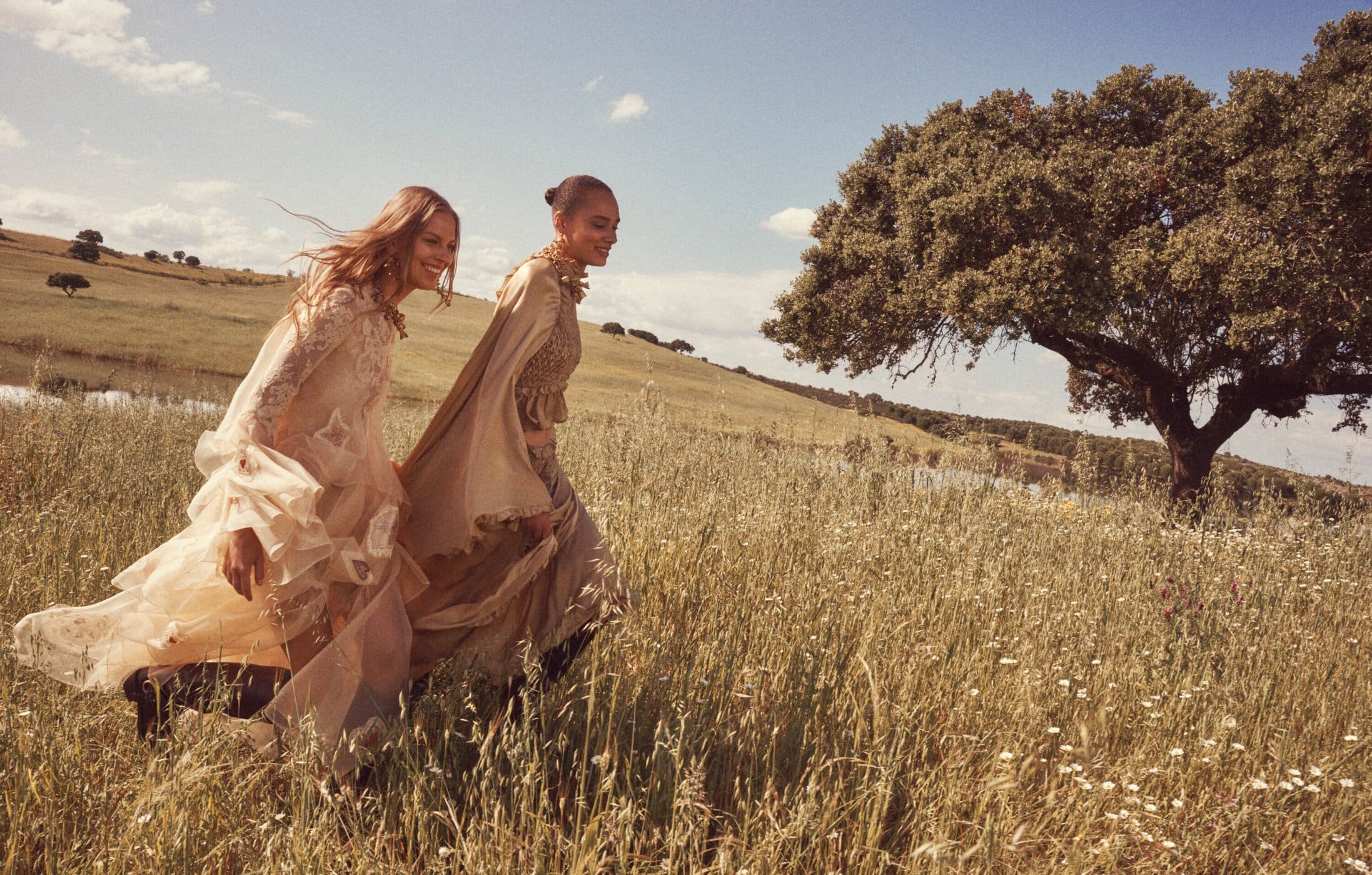 Two female models walking through a grassy meadow wearing flowing neutral-toned dresses under a blue sky