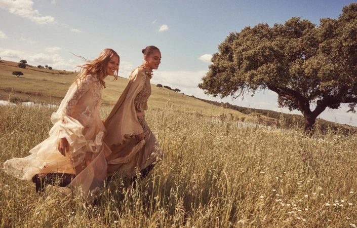 Two female models walking through a grassy meadow wearing flowing neutral-toned dresses under a blue sky