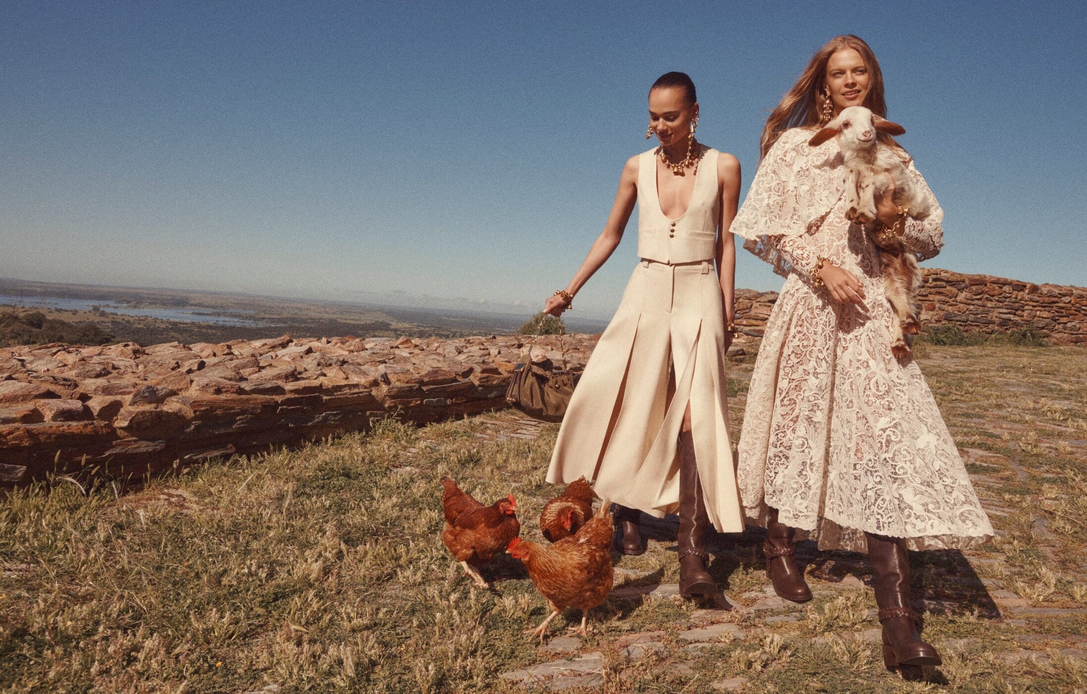 Two female models posing outdoors on a sunny hill with chickens and a lamb, photographed by Benny Horne for Zimmermann.