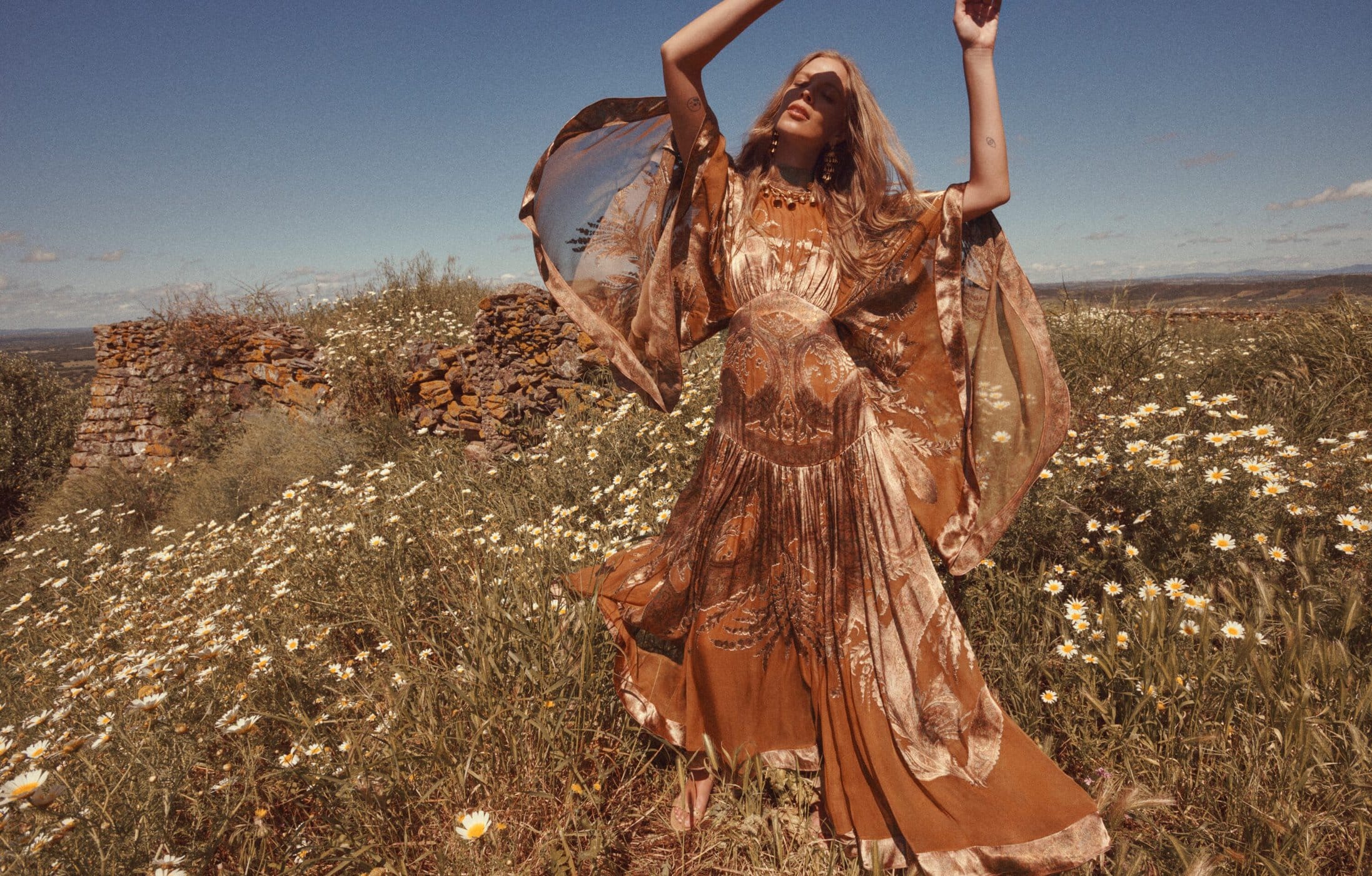 Model in a brown patterned dress standing in a wildflower field with arms raised under a blue sky