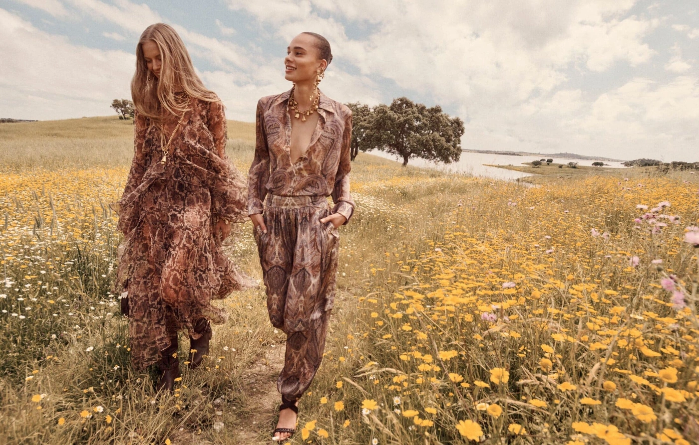 Two female models walking through a grassy field filled with yellow wildflowers under an open cloudy sky.