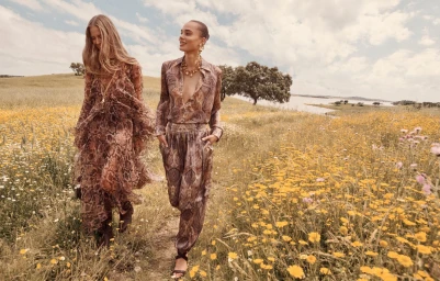 Two female models walking through a grassy field filled with yellow wildflowers under an open cloudy sky.