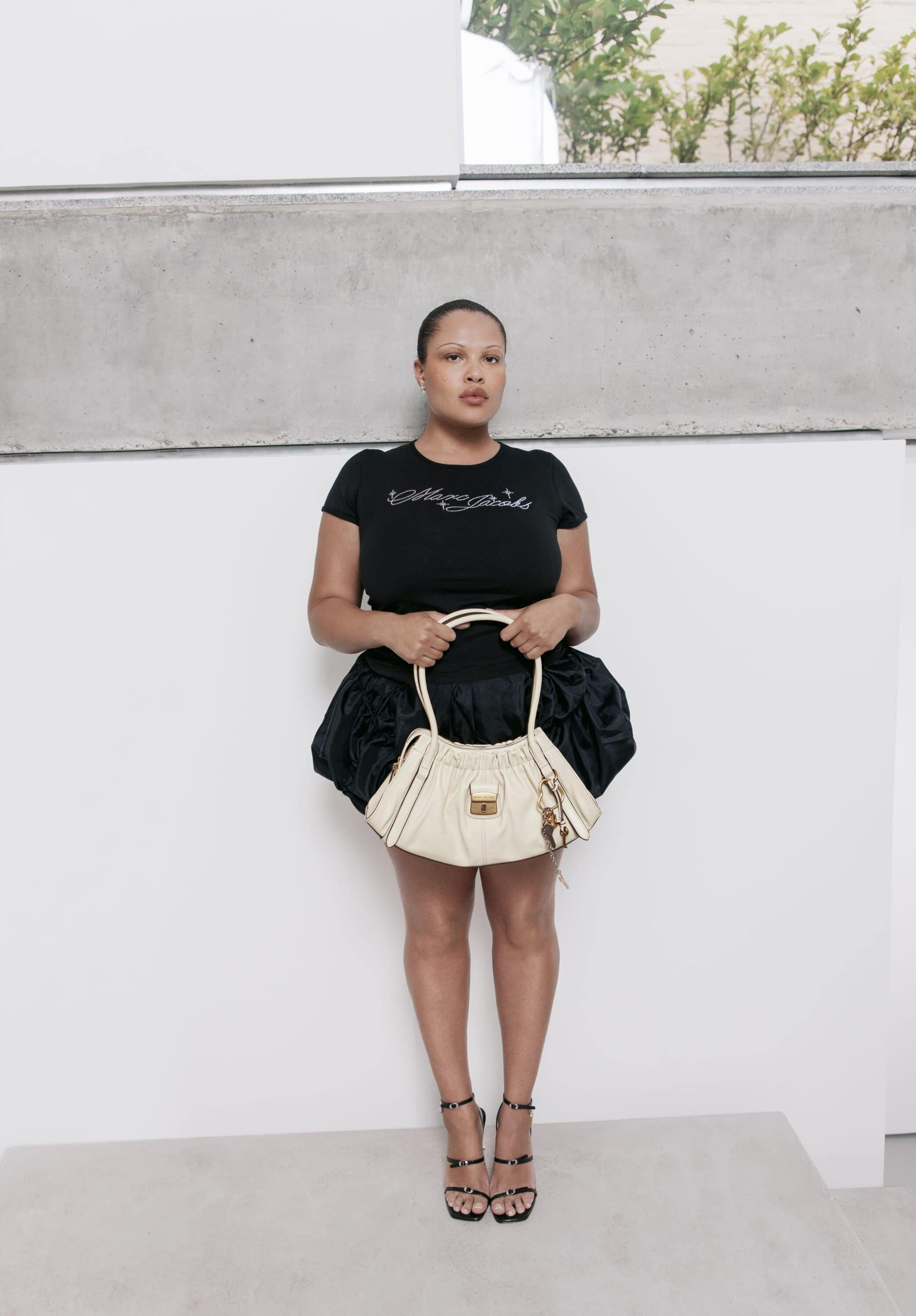 Model wearing a black graphic t-shirt and ruffled skirt holding a cream handbag against a plain white wall.