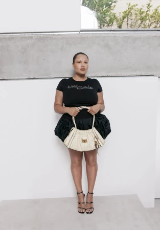 Model wearing a black graphic t-shirt and ruffled skirt holding a cream handbag against a plain white wall.