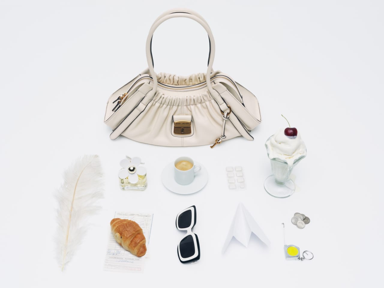 White leather handbag surrounded by arranged personal items like coffee, sunglasses, croissant, and dessert on white background