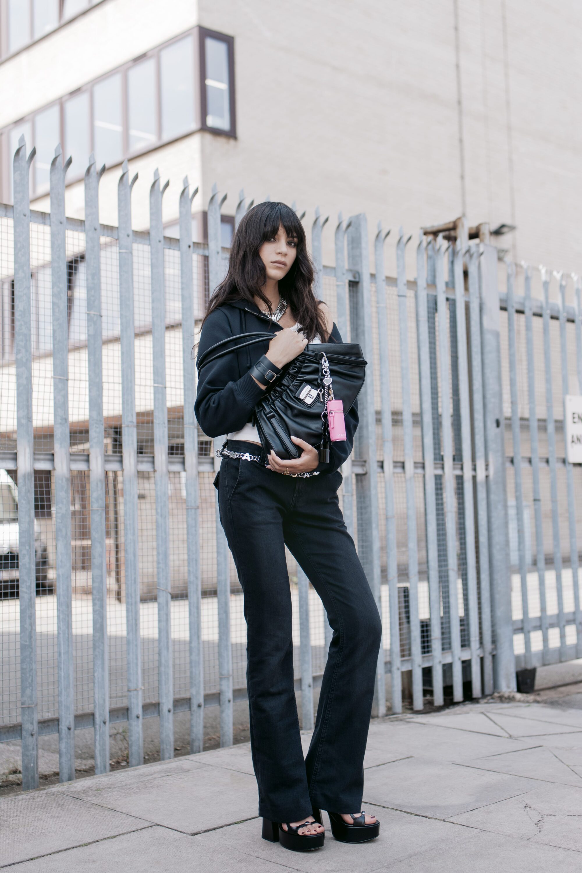 Model standing outdoors wearing a black zip hoodie, dark pants, and holding a large black leather handbag.