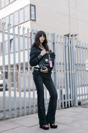 Model standing outdoors wearing a black zip hoodie, dark pants, and holding a large black leather handbag.