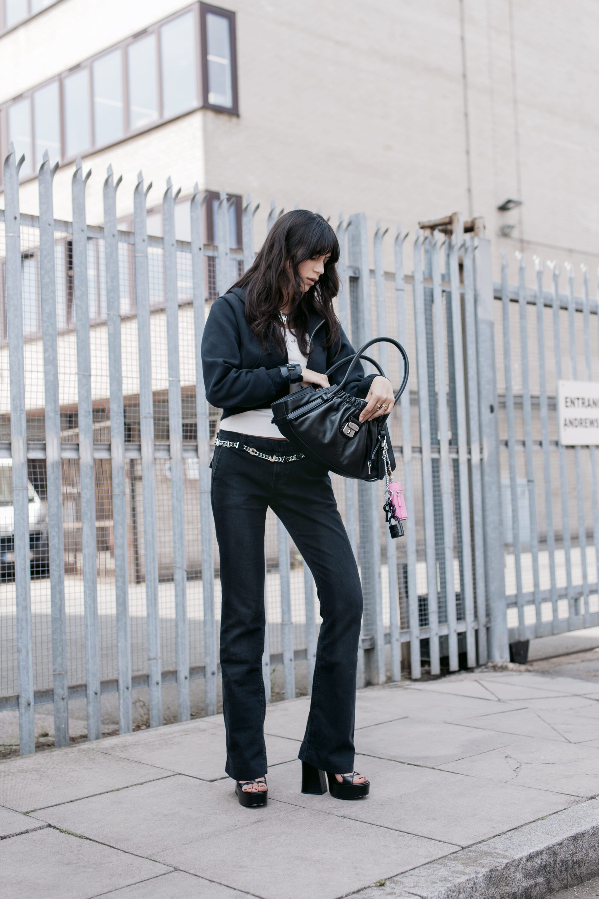Woman in dark zip hoodie and black trousers holding a black handbag standing outside a metal spiked fence.