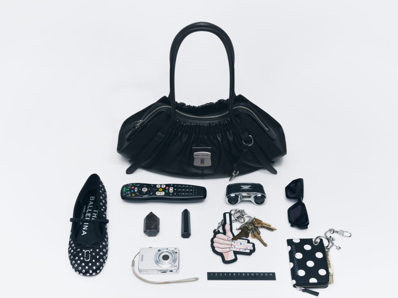 A black leather handbag displayed flat-lay style alongside accessories on a plain white background for Marc Jacobs