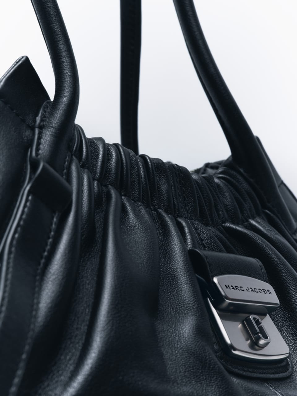Close-up view of a black leather handbag featuring a metallic buckle engraved with the Marc Jacobs logo against a white background.