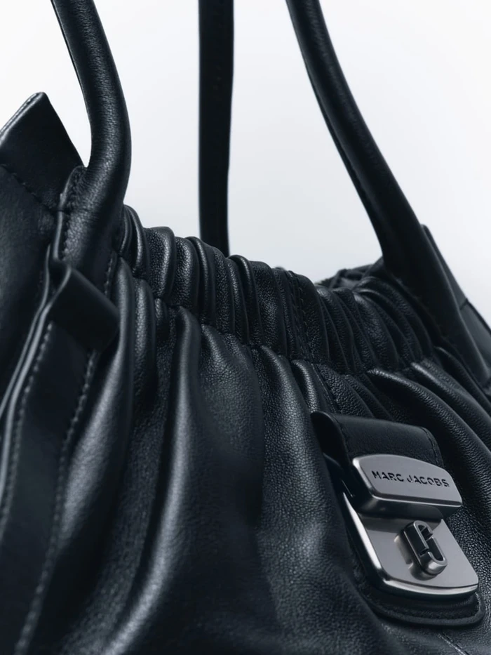 Close-up view of a black leather handbag featuring a metallic buckle engraved with the Marc Jacobs logo against a white background.