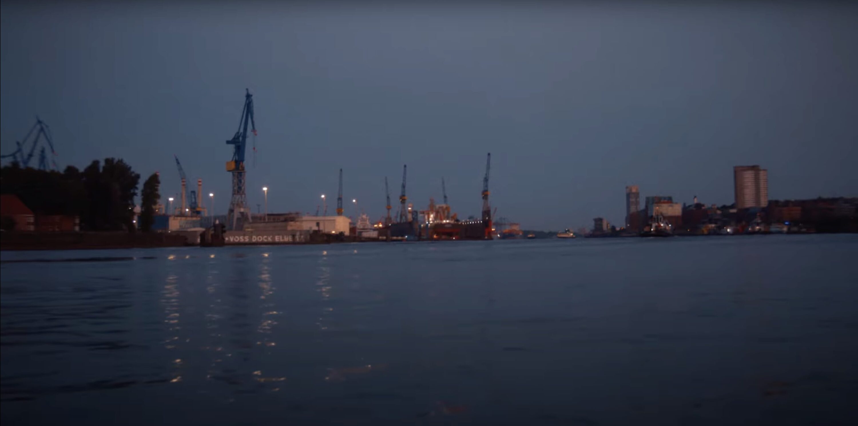 Dusk view across a dark river toward industrial docks with towering port cranes and distant city lights under an overcast sky.