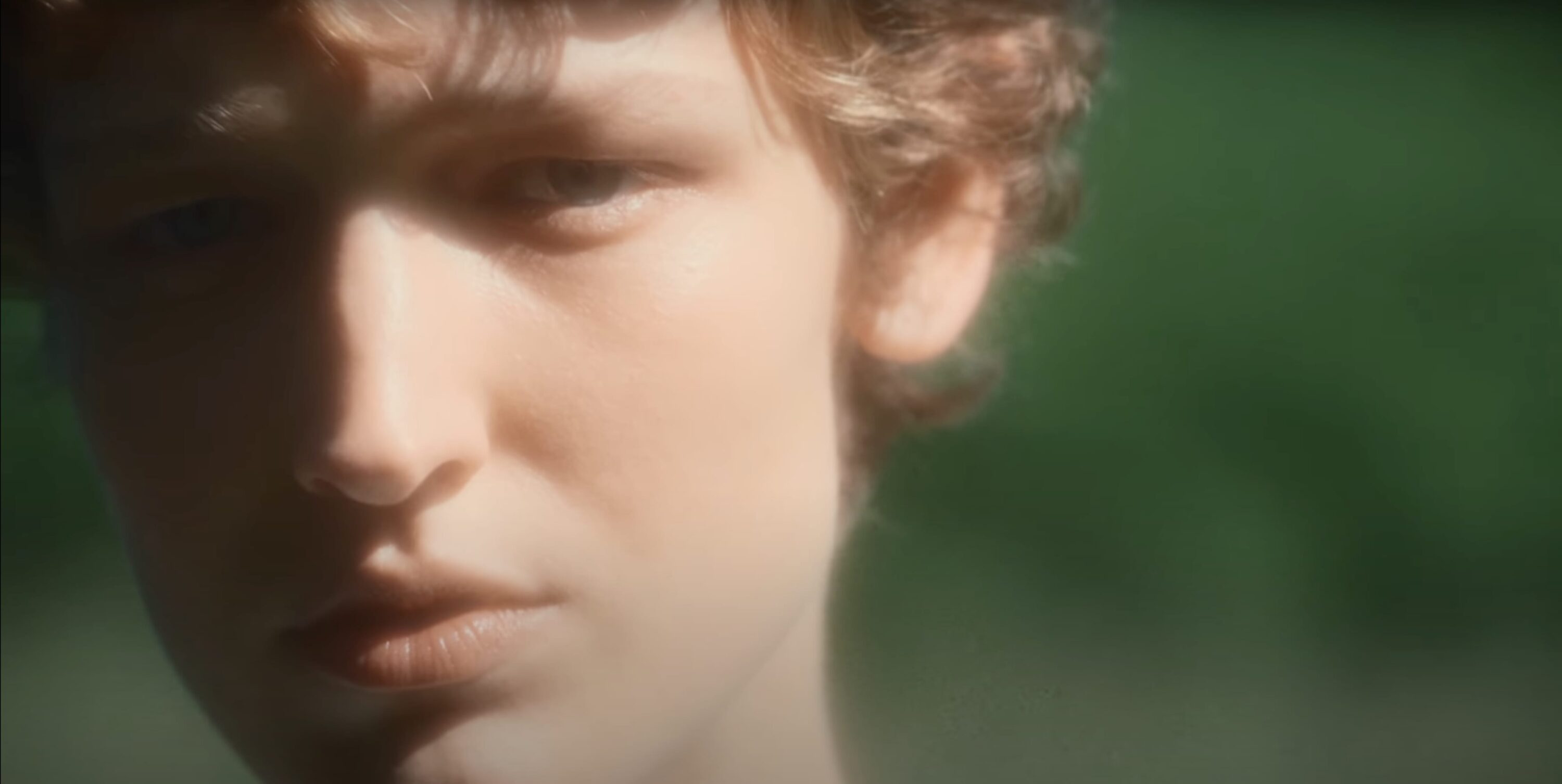 Close-up portrait of a young person with light curly hair outdoors against a green blurred background for Jil Sander.