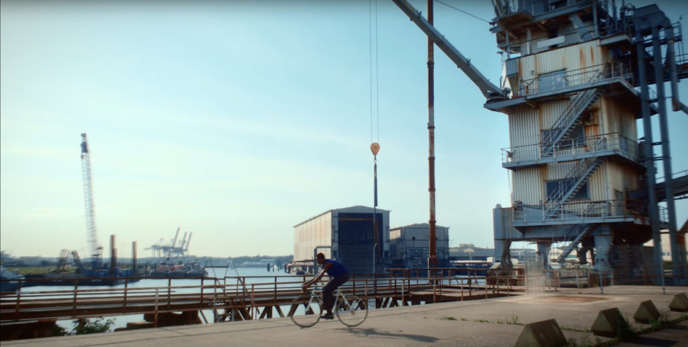 Man riding a bicycle on a wide paved dock near industrial cranes and a large building with stairs under a clear sky.
