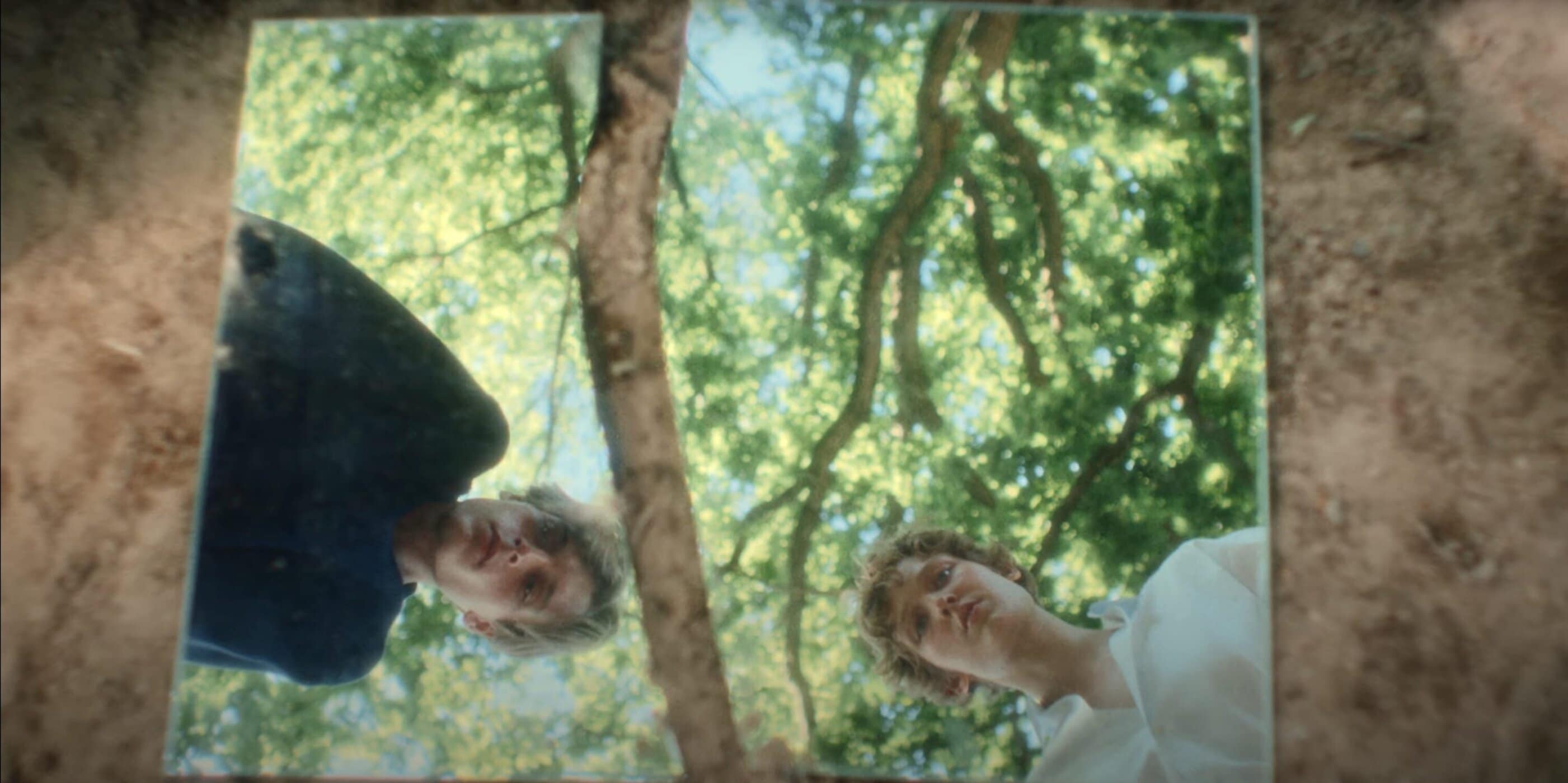 Two people reflected in mirror shards on dirt ground showing overhead trees