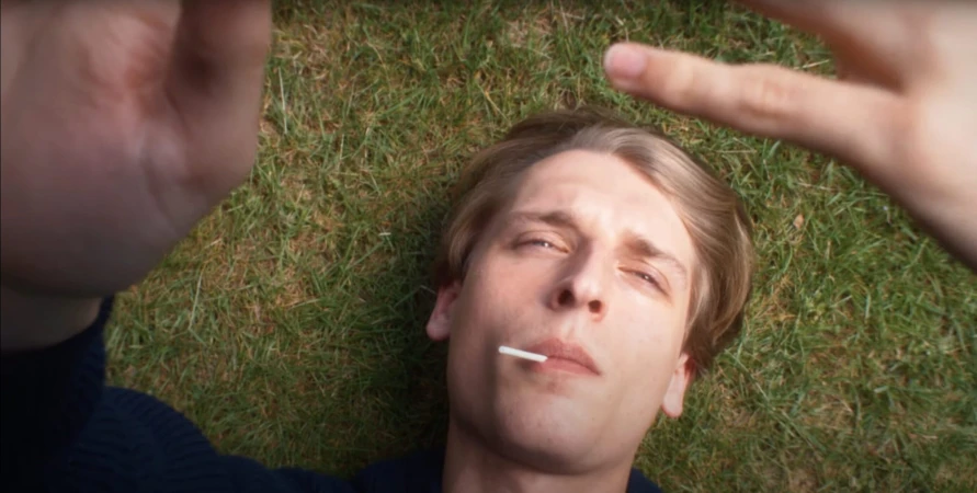 A young man lying on green grass looking upward with hands raised in the foreground, for Jil Sander.