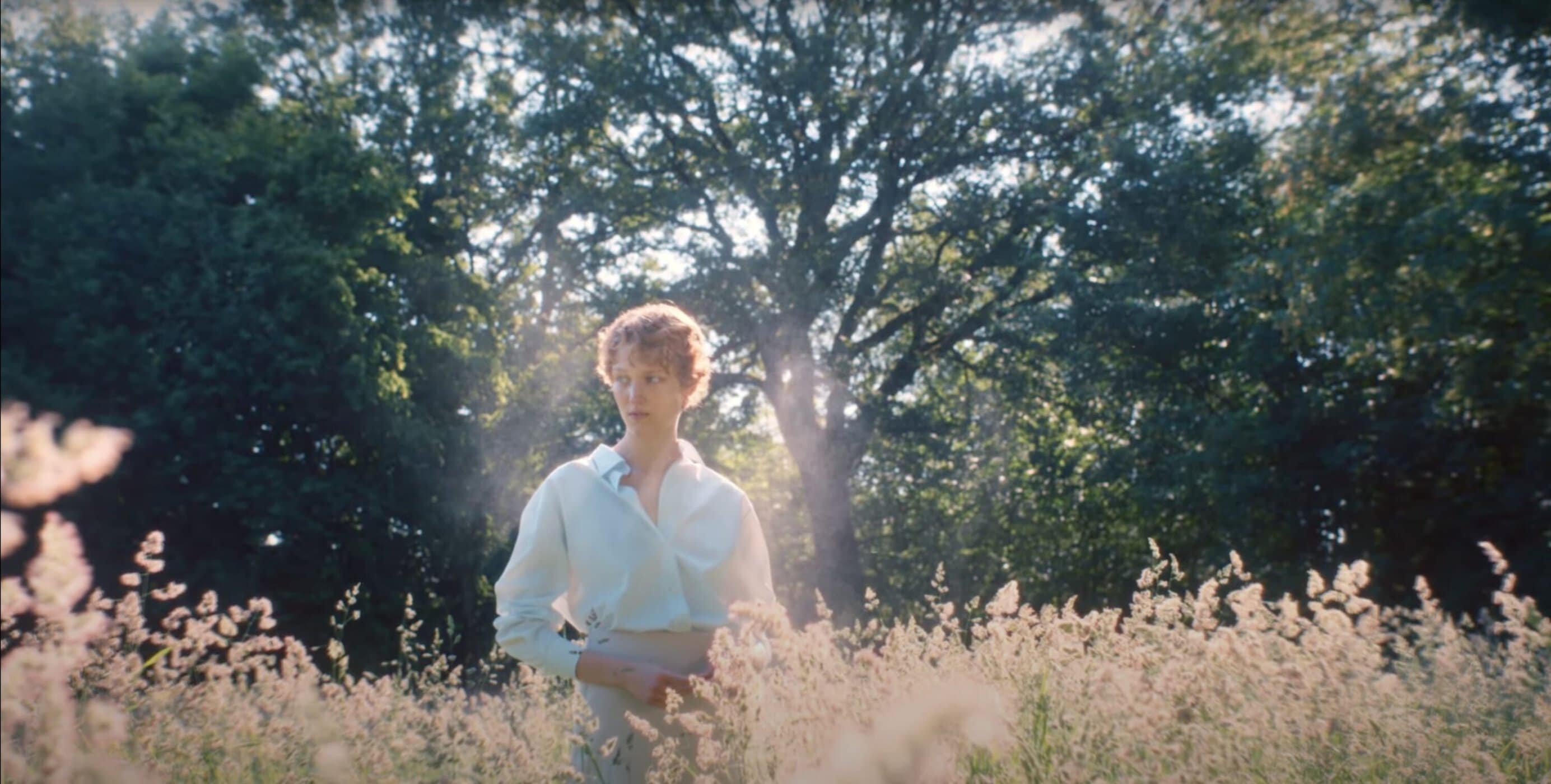 Woman in a light shirt standing amidst tall grass in a sunlit forest clearing for Jil Sander Wanderlust ad campaign.