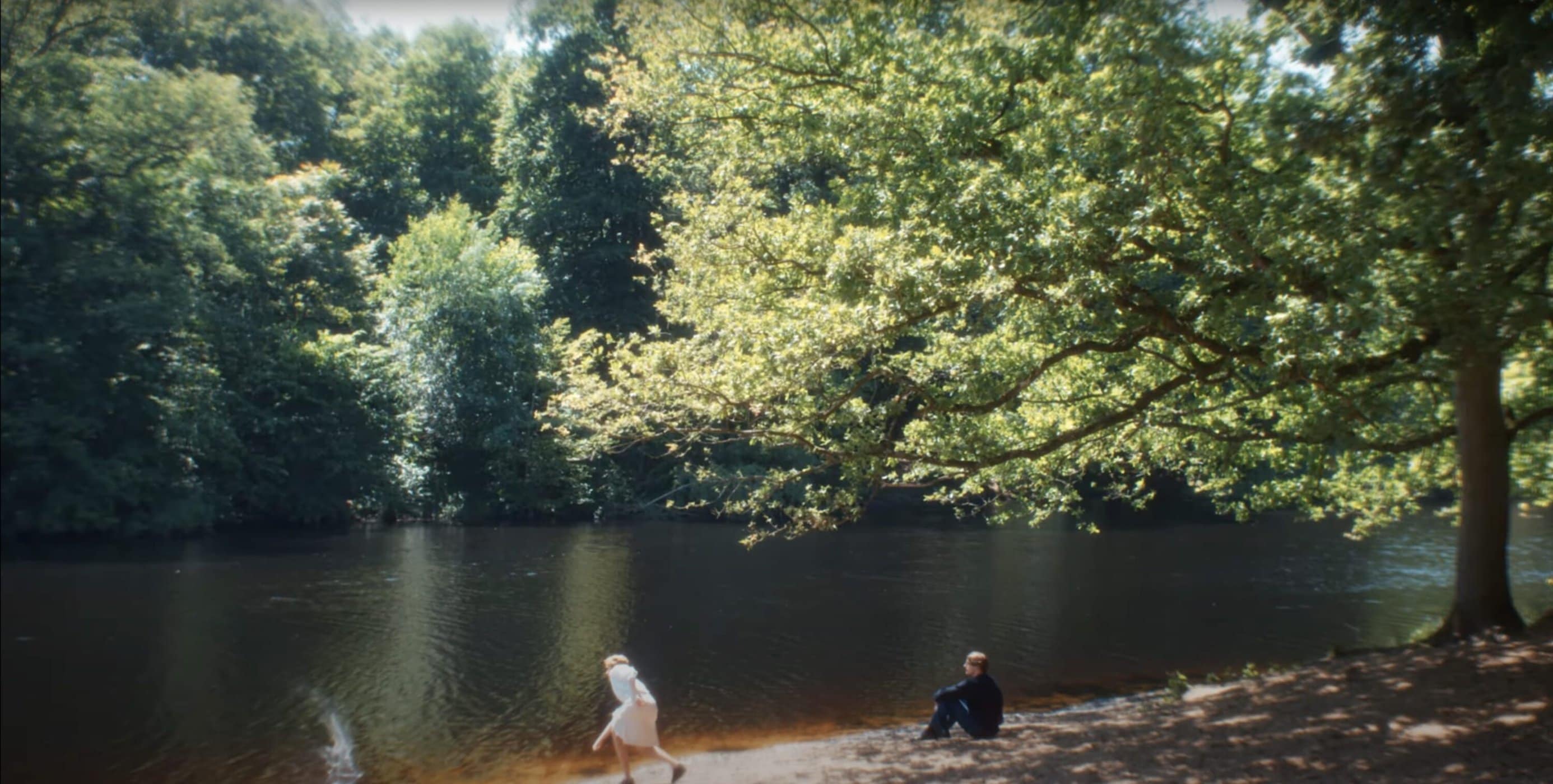 Two people by a riverbank in a lush forest, one standing and one seated near large overhanging tree branches.