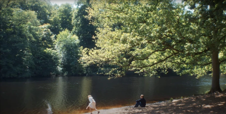 Two people by a riverbank in a lush forest, one standing and one seated near large overhanging tree branches.