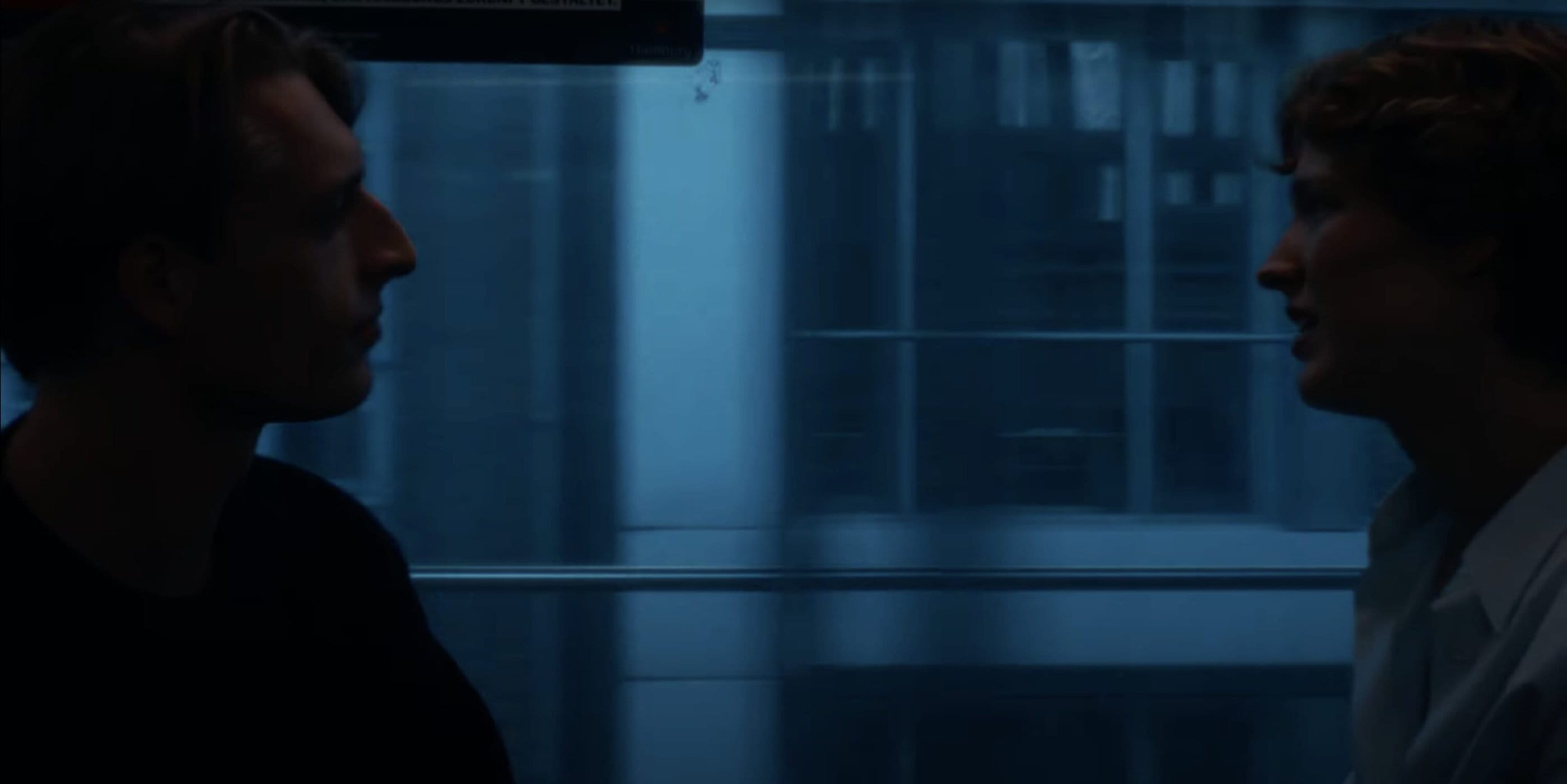 Two people in profile facing each other inside a train car with a blue-toned city backdrop visible through the window.
