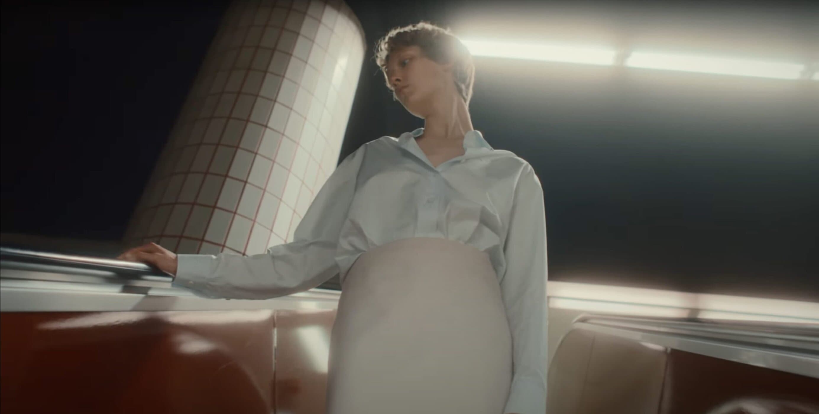 Model in light blue shirt standing indoors next to a tiled pillar with warm overhead lighting for Jil Sander.