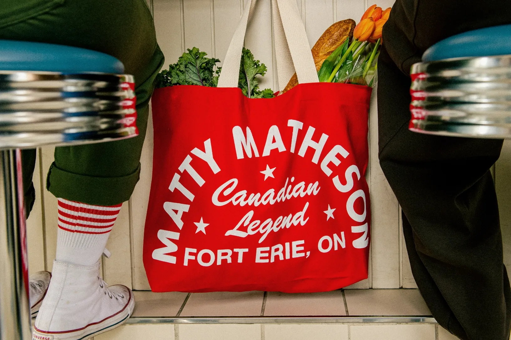 Red canvas tote bag with white text filled with groceries and flowers, flanked by two people seated on stools.