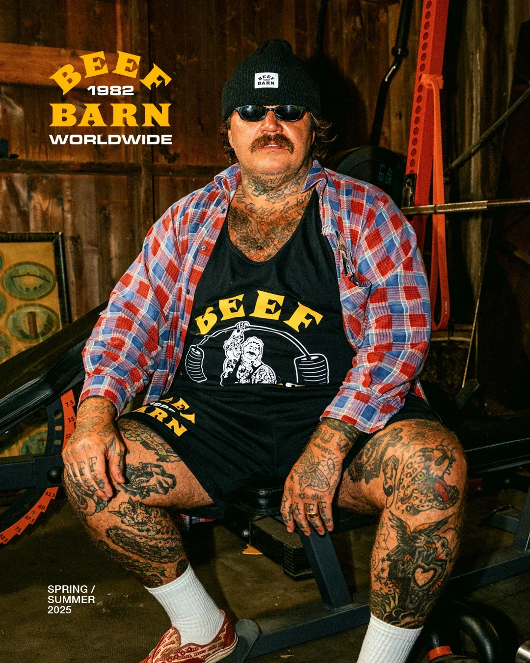 Man wearing a black beanie and sunglasses sitting in a gym wearing a plaid shirt and black shorts with graphic text