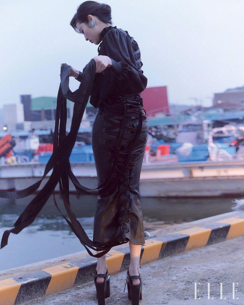 High-fashion editorial shot of a slender woman with dark hair on a pier, wearing a black dress and platform heels with boats in the background.