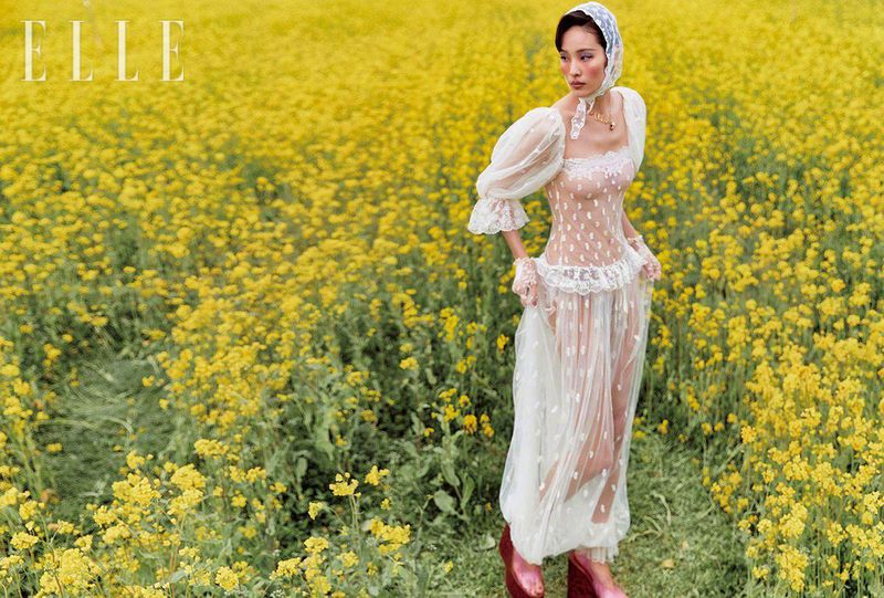 An Asian woman in a sheer white dress and headscarf poses in a field of bright yellow flowers for an outdoor fashion editorial.