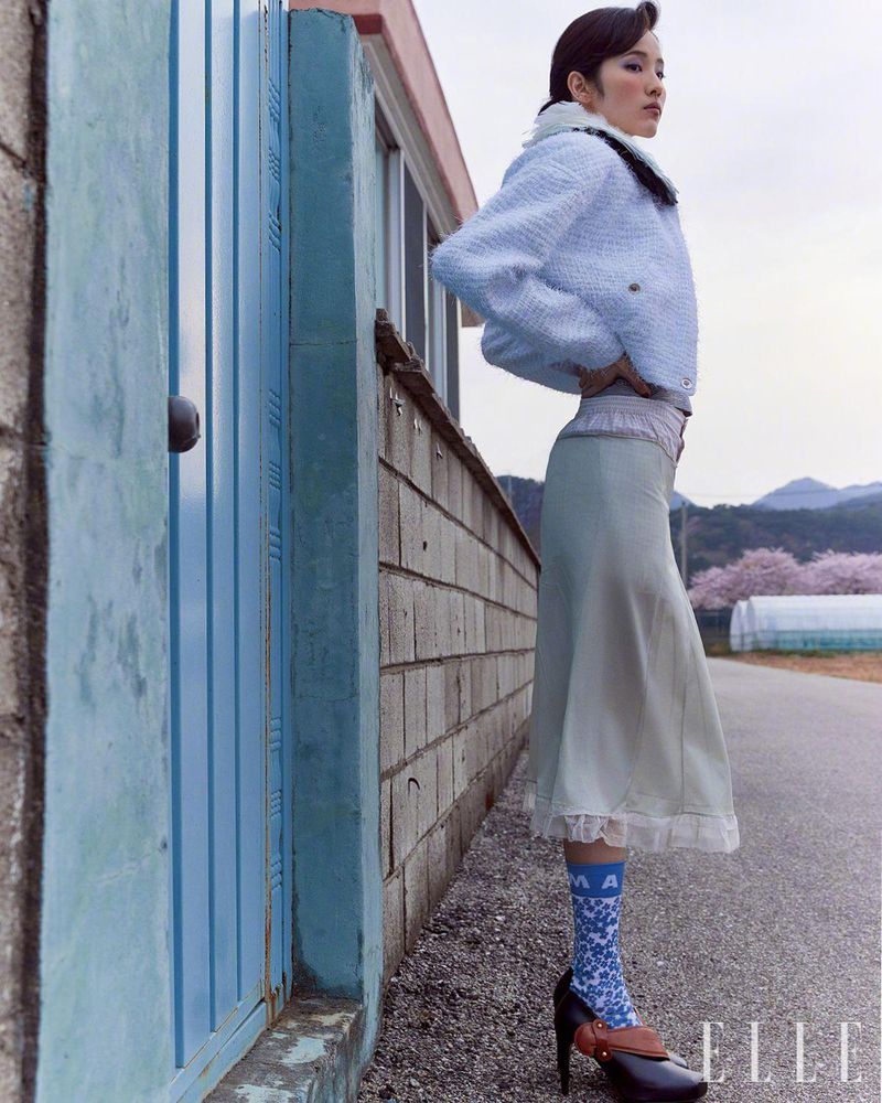 A stylish Asian woman poses in a light blue jacket and long skirt on a rural road with mountains and cherry blossoms in the background.