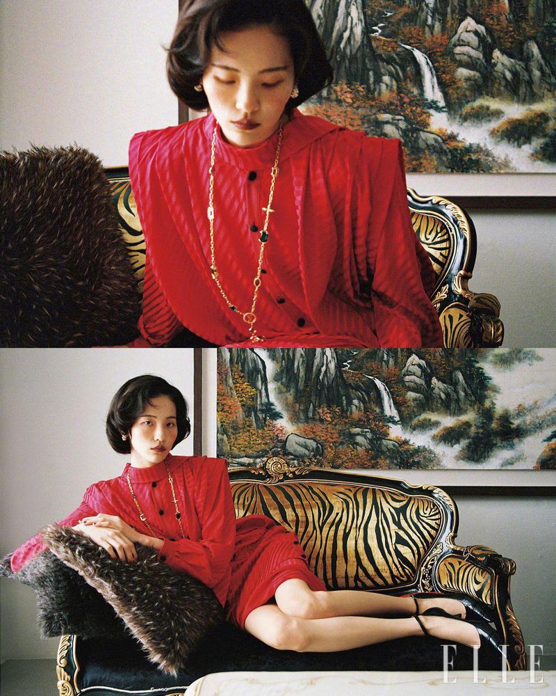 A woman in a red dress poses on a patterned couch near a nature painting, highlighted by classic fashion and elegant decor.