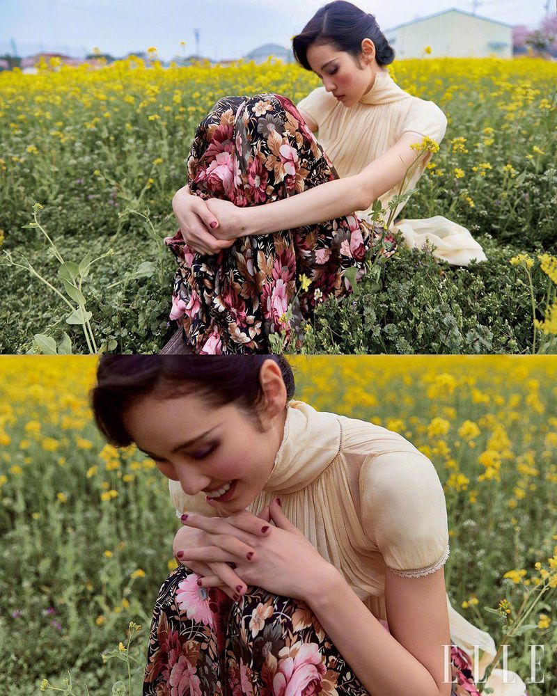Woman in floral dress sits in a yellow wildflower field, looking down. Outdoor fashion shot with natural light and soft focus.