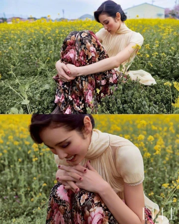 Woman in floral dress sits in a yellow wildflower field, looking down. Outdoor fashion shot with natural light and soft focus.
