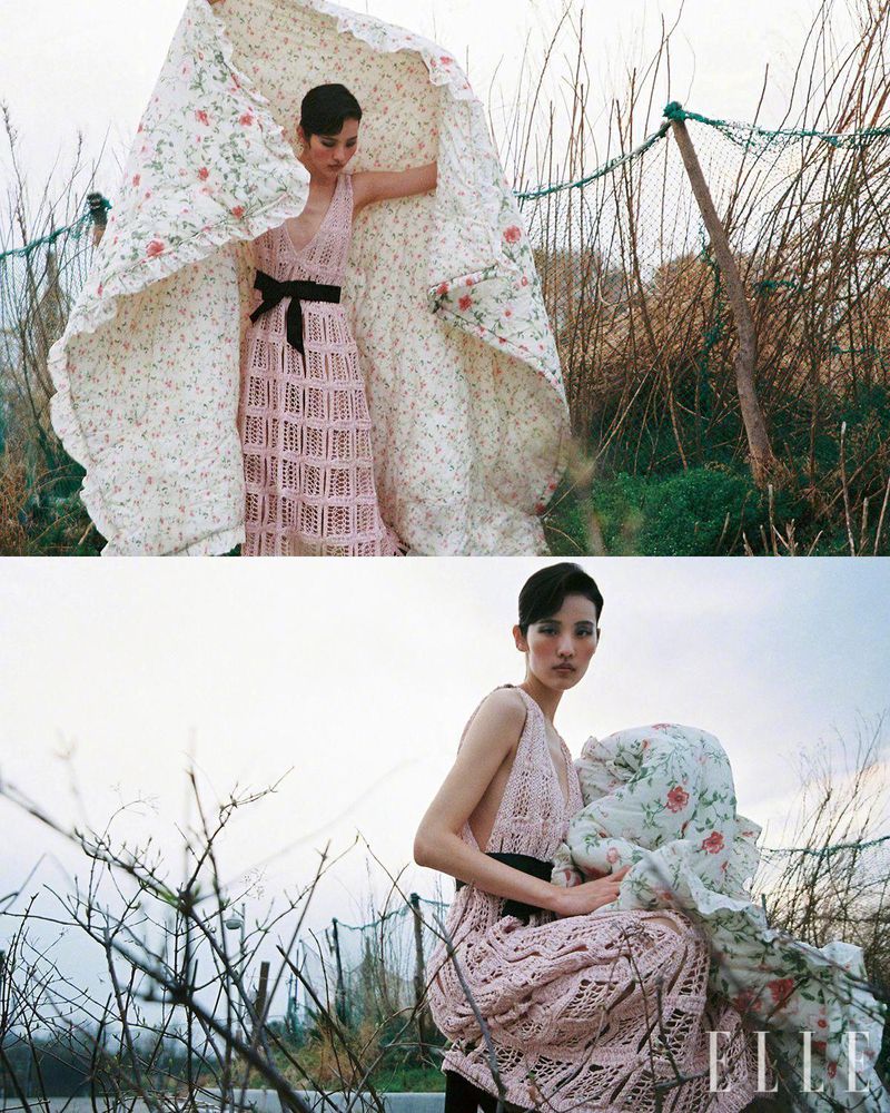 An Asian woman poses in a pink dress outdoors, holding a floral quilt with a blurred background of brush and foliage, fashion shoot.
