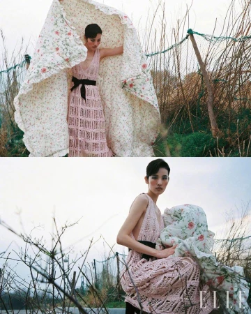 An Asian woman poses in a pink dress outdoors, holding a floral quilt with a blurred background of brush and foliage, fashion shoot.