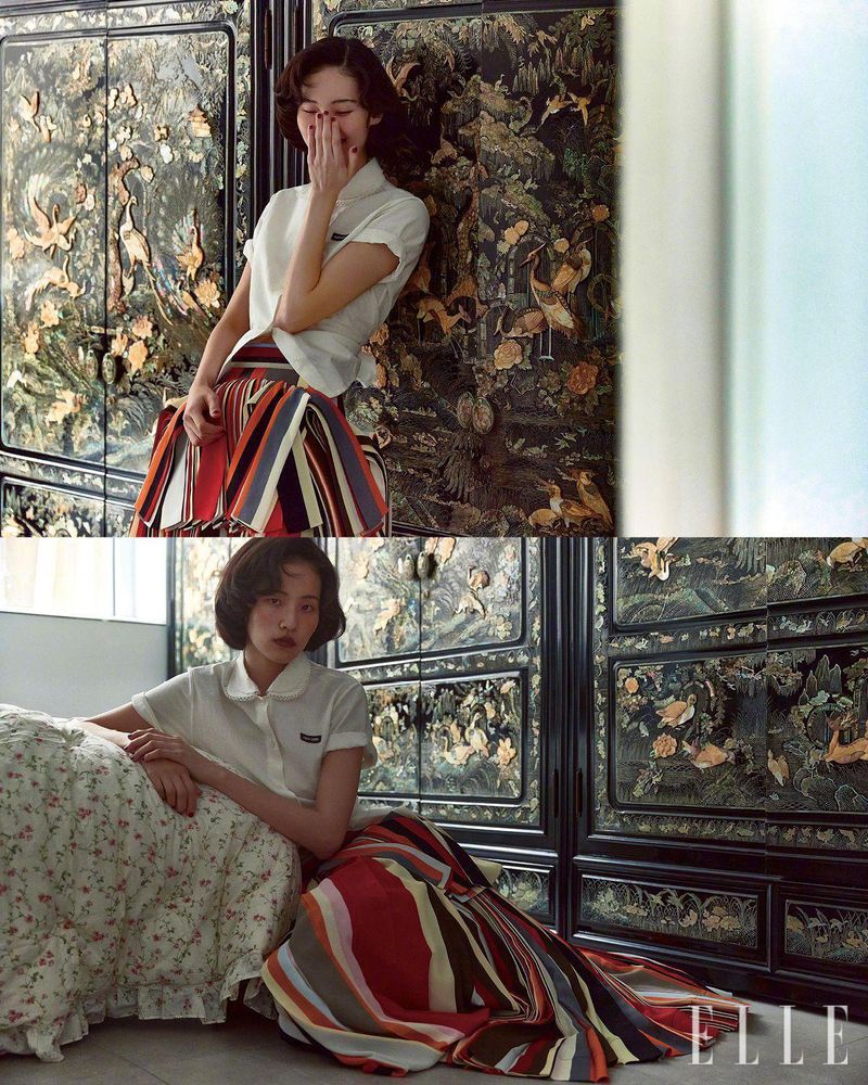 A young woman poses in a fashion shot, wearing a collared shirt and striped skirt. She is leaning against a carved cabinet.