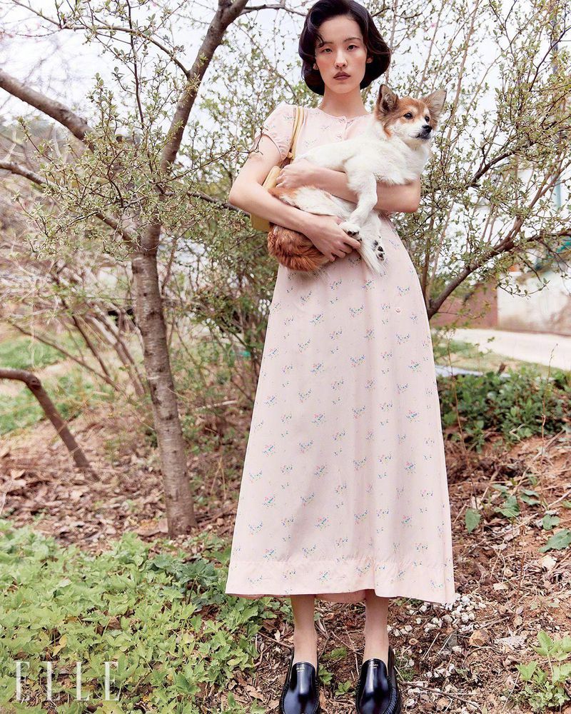 An Asian woman poses outdoors in a floral dress, holding a fluffy dog, with trees and a building in the background on a cloudy day.