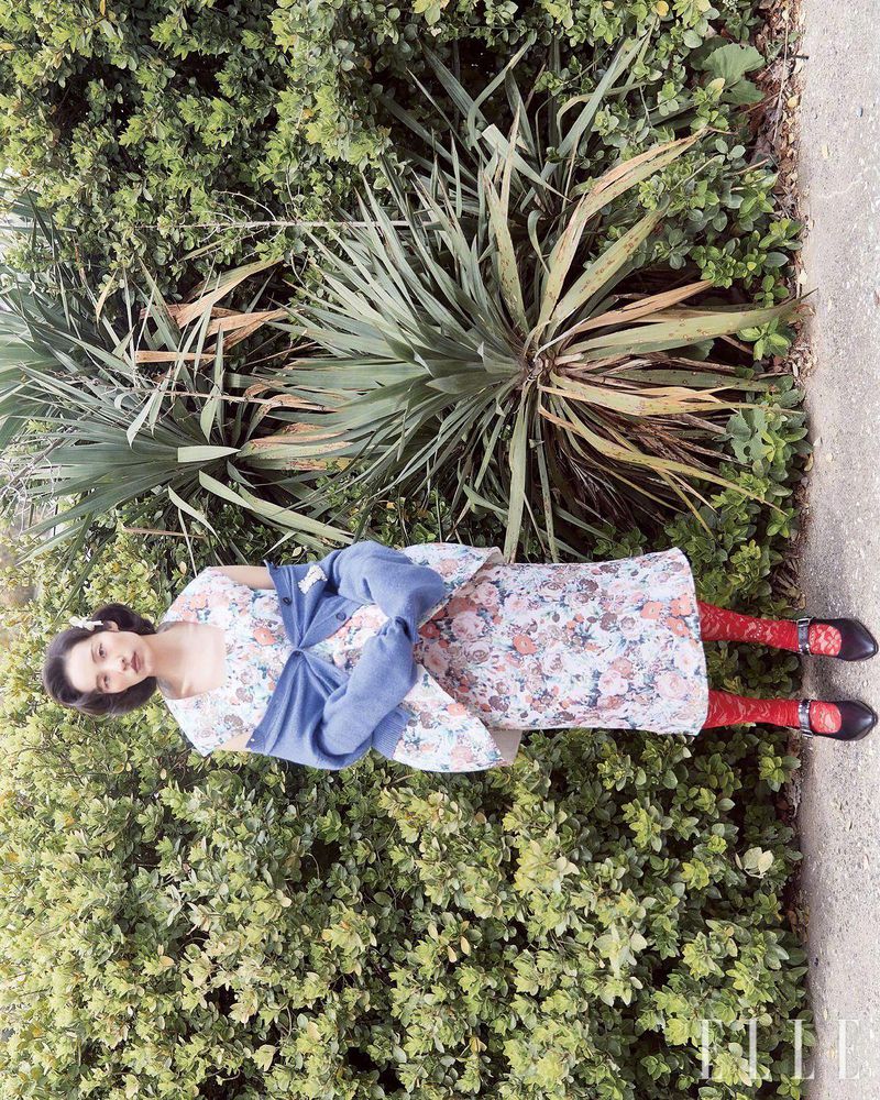 Adult female model in floral dress and red tights poses in lush green outdoor setting with blue jacket on dress and dress shoes.