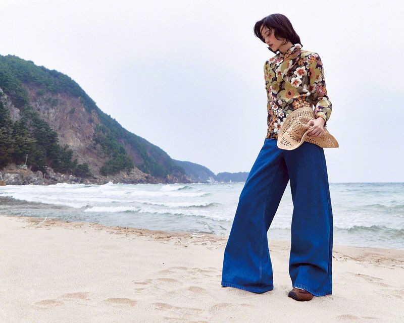 Woman posing on beach in floral shirt and wide-leg jeans, holding straw hat. Coastal scene with overcast sky and distant cliffs.