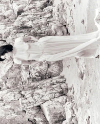 Model in a long white dress posing against a textured rocky cliffside background