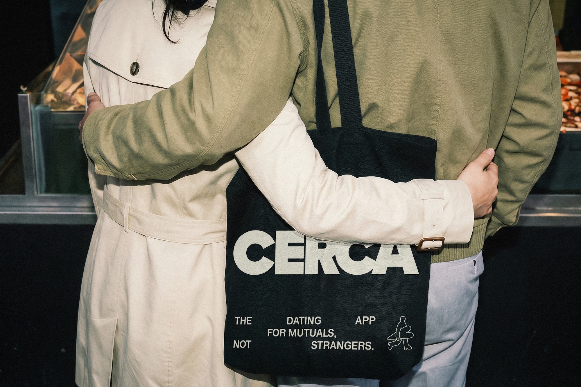 Two people embracing while wearing outerwear, holding a black canvas tote bag with CERCA printed on it.