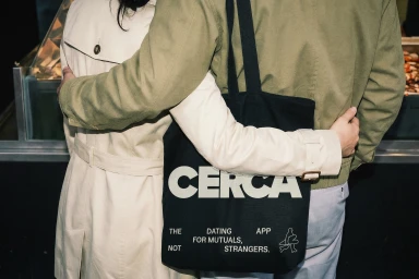 Two people embracing while wearing outerwear, holding a black canvas tote bag with CERCA printed on it.